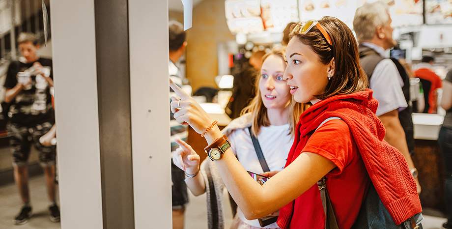 Two young women interact with a self-service kiosk touchscreen at a fast-service restaurant